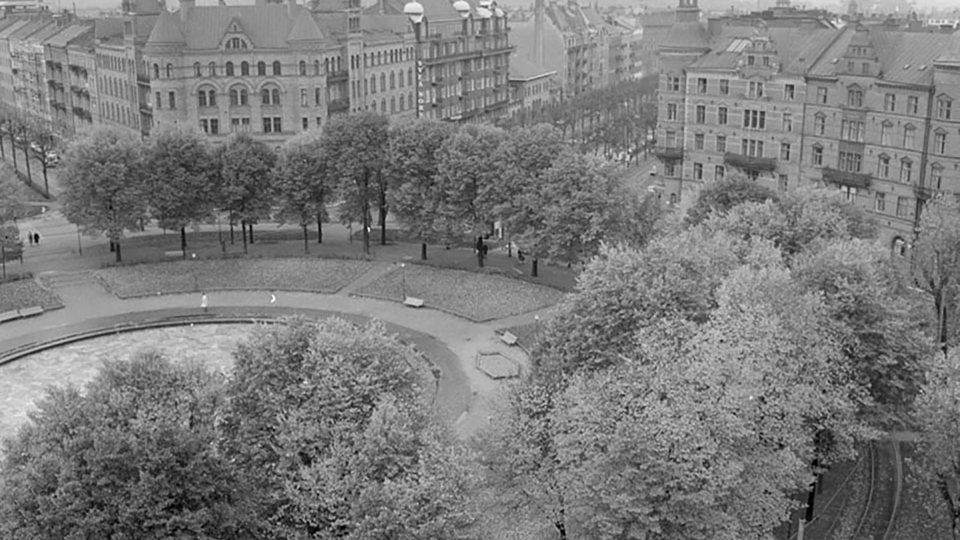 Vy över Karlaplan 1960 med gräsytor och gångvägar, omgiven av träd och hus, svartvitt foto.